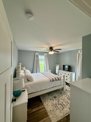 Une chambre à coucher avec un lit, un ventilateur de plafond, une commode, un téléviseur et une fenêtre avec des rideaux.