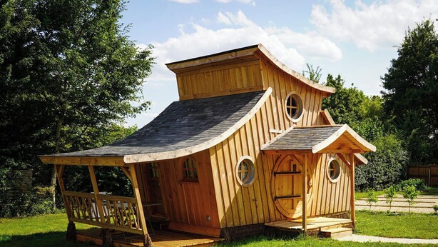 A wooden house with a sloped roof, circular windows, and a porch. - The Hobbit House at the Crossroads (Fontenoy)