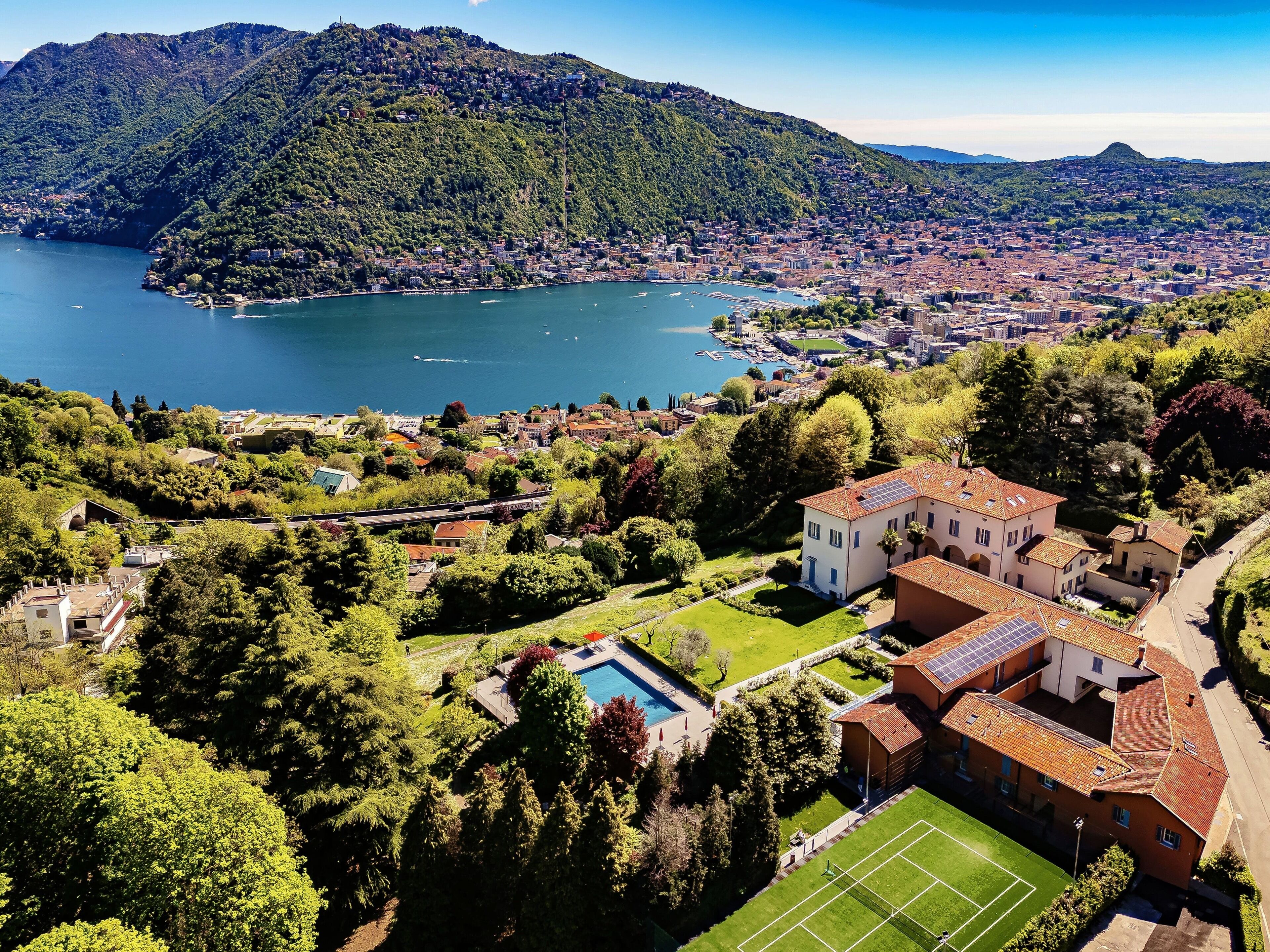 A residential area with a large house featuring a swimming pool and solar panels, surrounded by lush greenery and a lake.