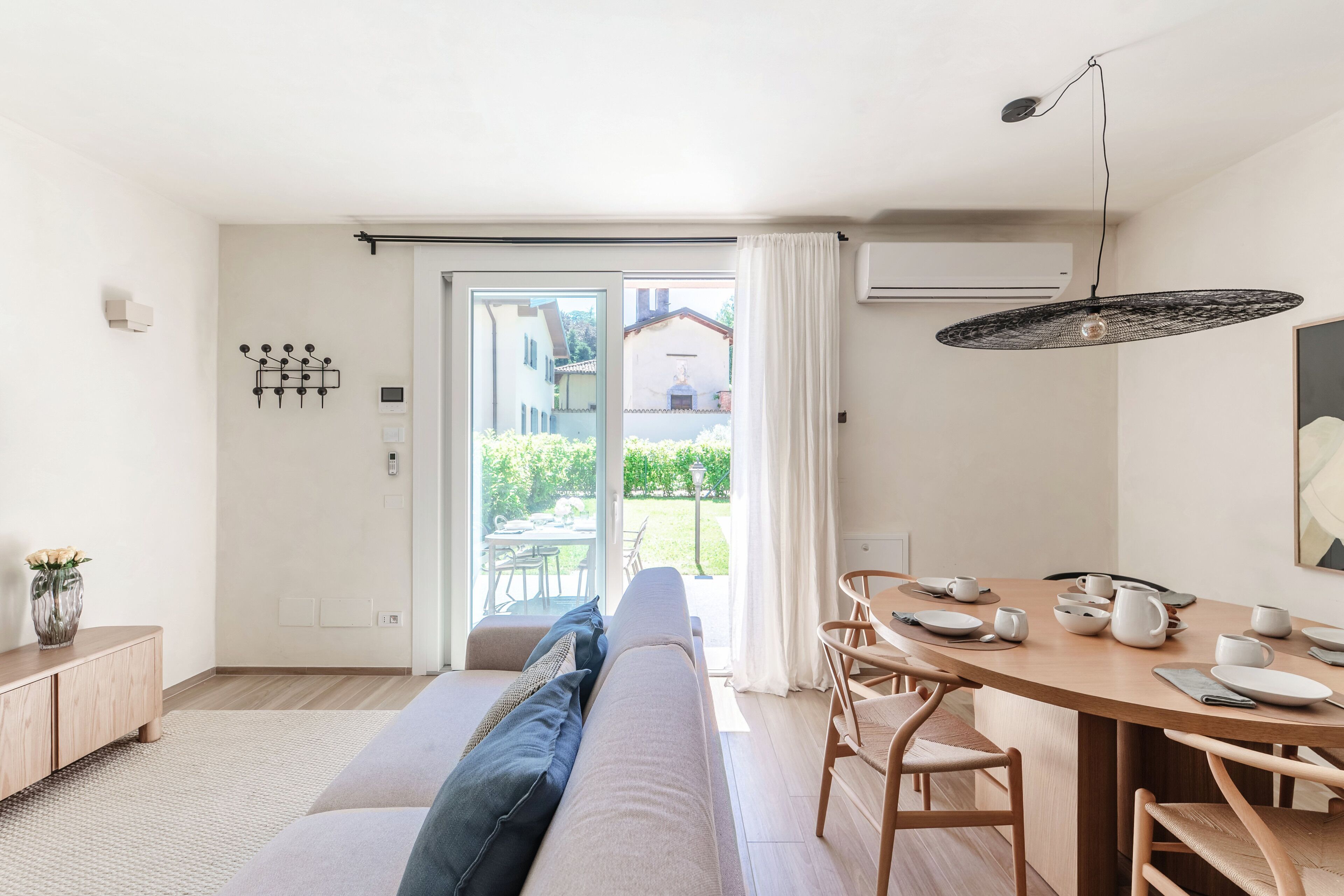 Una sala moderna con sofá, mesa de comedor y sillas, un ventanal corredizo a un jardín, y una lámpara colgante.