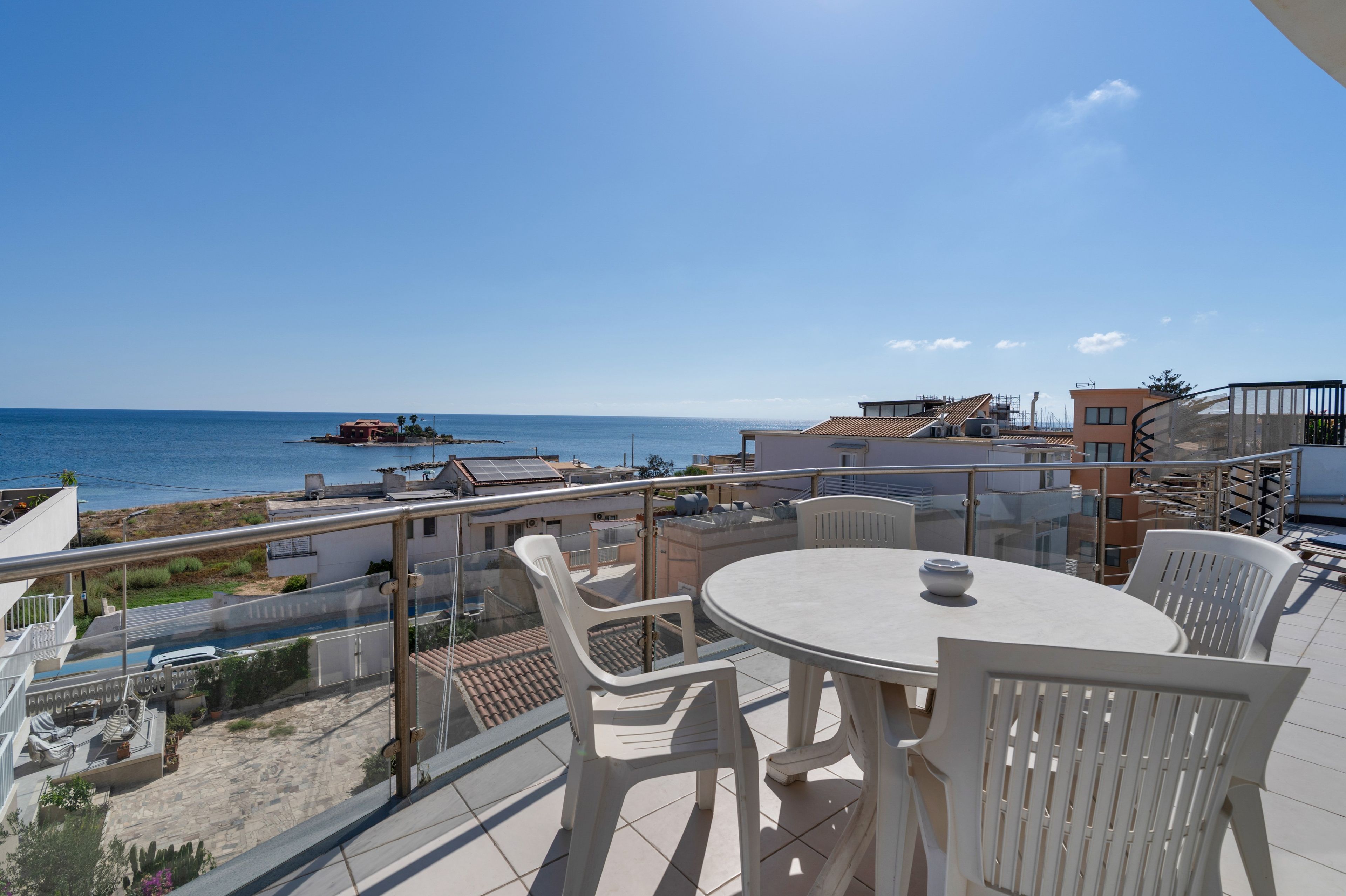 A balcony with a table and chairs, overlooking a coastal area with buildings and a body of water.