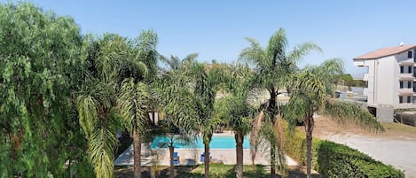 Zona de piscina con una piscina azul cristalina, rodeada de árboles y un edificio con tejado de azulejos.