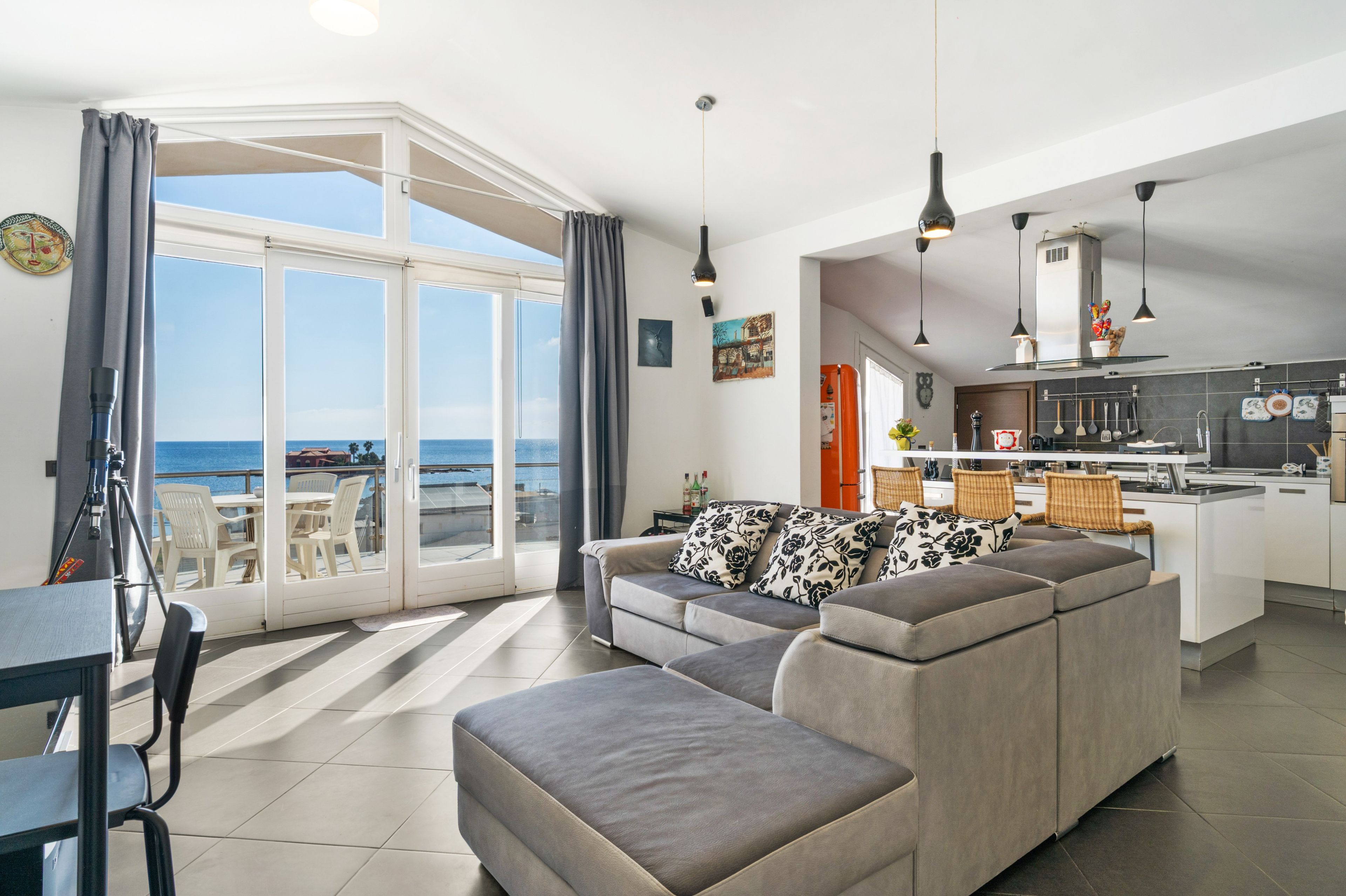 A modern living room with a grey sofa, a dining area with a table and chairs, and a kitchen with hanging lights.