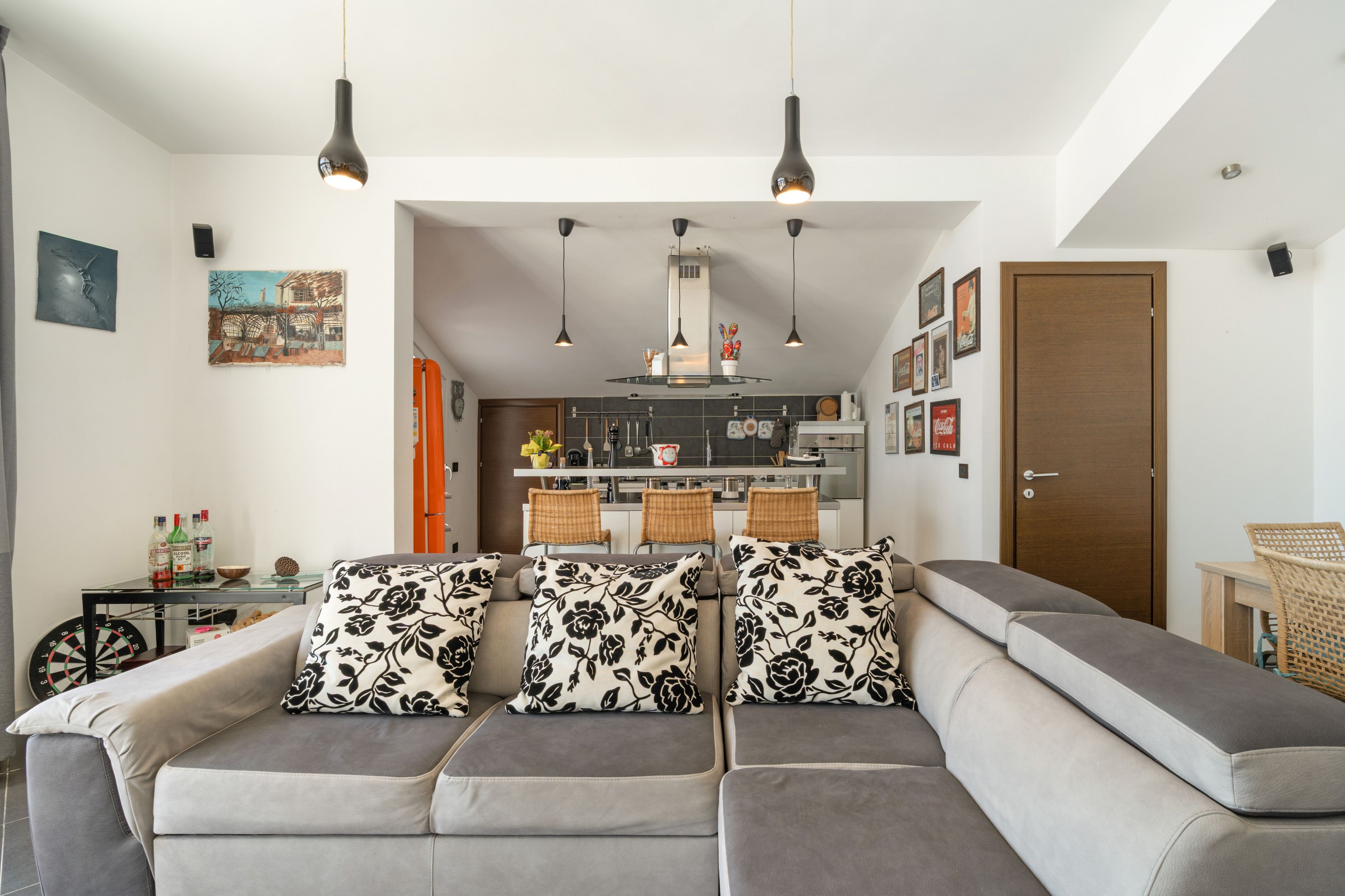 Ein modernes Wohnzimmer mit einem grauen Sofa, Kissen in Schwarzweiß-Muster und einer Küche im Hintergrund.