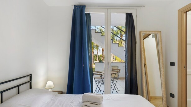 A bedroom with a bed, a nightstand, a mirror, and a view of an outdoor area with a table and chairs. - Casa Marea Ground Floor with Garden, Wi-Fi and Air Conditioning (Marzamemi)