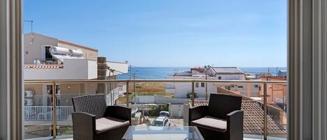 A balcony with wicker furniture, a glass table, and a view of the sea and buildings.