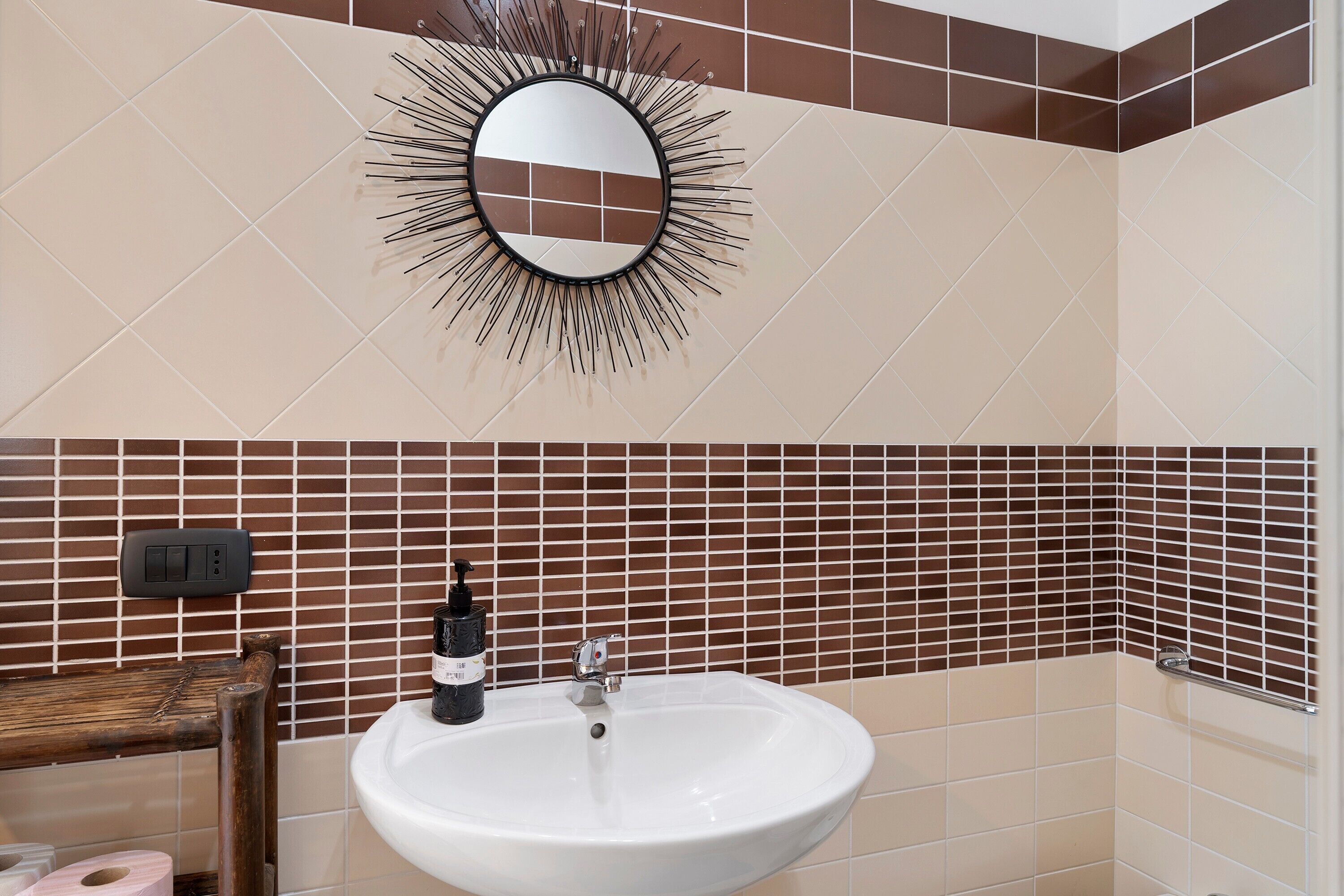 Ein Badezimmer mit einem weißen Waschbecken, einem Spiegel mit Sonnendesign und braunen sowie beige gefliesten Wänden.