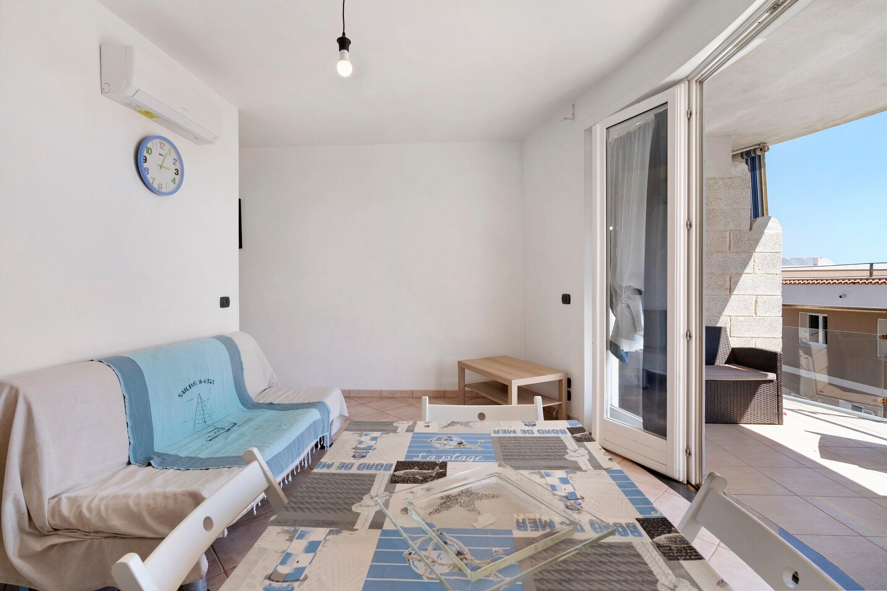 A room with a sofa, a small table, a glass door leading to a balcony, and a wall clock.