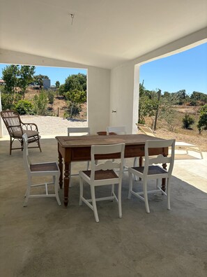 An outdoor patio with a wooden table and white chairs, a wicker chair, and a view of a hilly landscape. - Country House 'Quinta Da Corte' with Private Garden, Balcony and Wi-Fi (Melides)