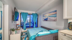 Un dormitorio con una cama, televisión, ventana con cortinas, un cuadro y una mesita con una planta.
