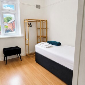 A small room with a bed, a black bench, a wooden wardrobe, and a window with a view of a brick building. - 3BR Hastings House | Contractor Friendly (Sunderland)
