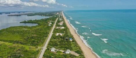 An aerial view of a coastal road with houses along the beach, a clear sky, and calm sea.