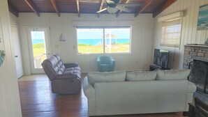 Ein geräumiges Wohnzimmer mit Holzboden, einem großen Fenster mit Blick auf den Strand, einem grauen Sofa, einem blauen Sessel und einem Kamin.