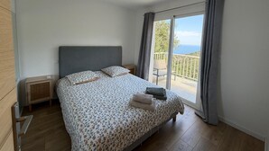 A bedroom with a bed, a nightstand, a window with curtains, and a view of the outdoors. - Charming apartment in the hills above Erbalunga (Brando)