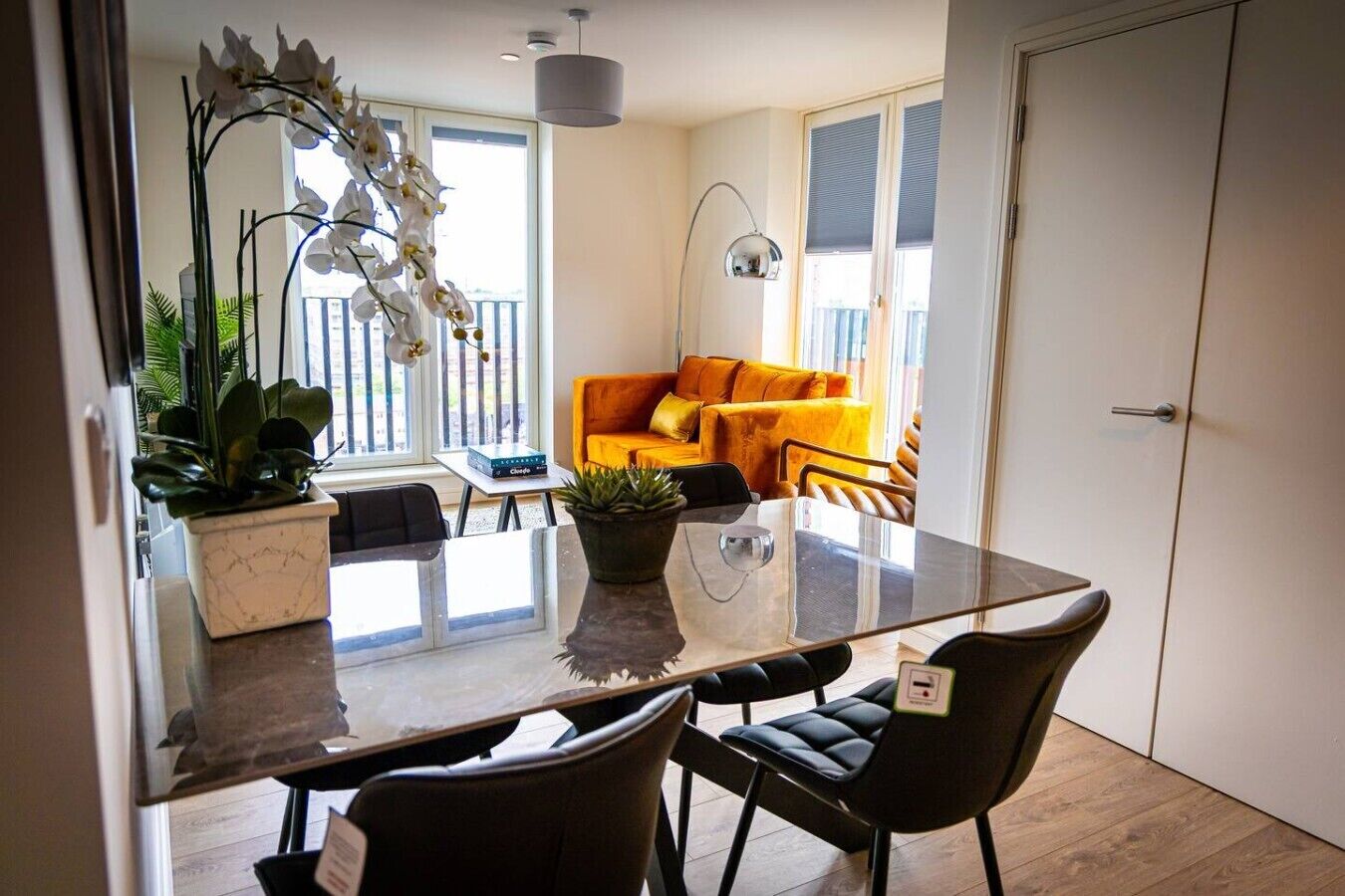 A modern dining area with a glass table, black chairs, a potted plant, and a sofa with cushions.