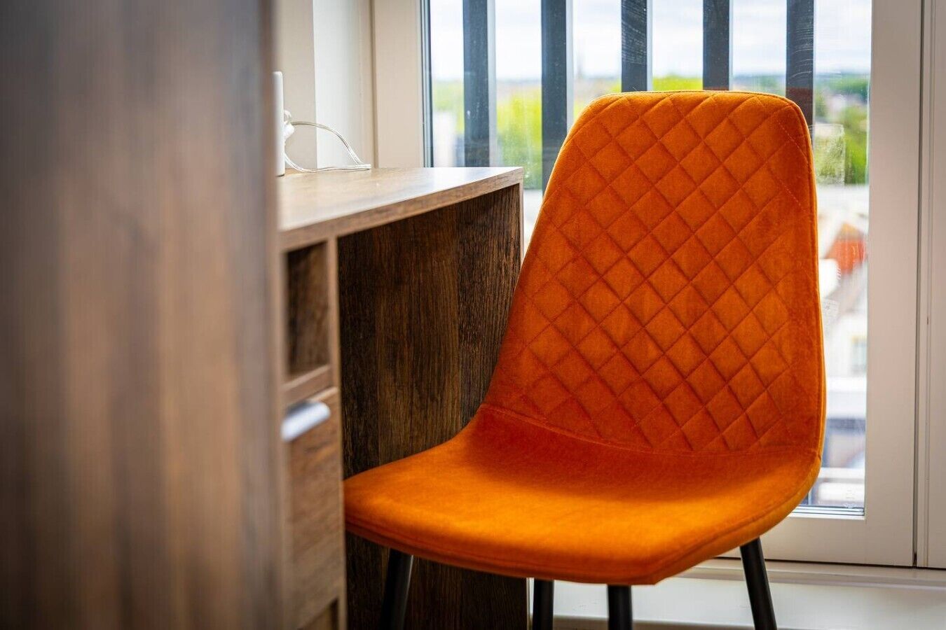 A chair with a quilted backrest and legs, positioned next to a wooden desk and a window with a view of greenery.