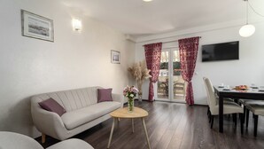 Ein Wohnzimmer mit einer Couch, einem kleinen runden Tisch mit einer Vase, einem Esstisch mit Stühlen, einem Fernseher und einem Fenster mit Vorhängen.