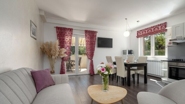 Ein modernes Wohnzimmer mit einer Couch, einem runden Holztisch mit Blumen, einem Essbereich mit Stühlen, einer Küche mit Schränken und Geräten sowie einem an der Wand montierten Fernseher.