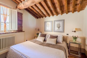 Un dormitorio con una cama, dos mesitas de noche, una ventana con cortinas, un radiador y dos cuadros en la pared.