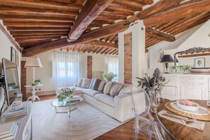 Una sala de estar estilo loft con vigas de madera a la vista, un sofá blanco, una mesa de centro de vidrio y una zona de comedor con una mesa redonda.