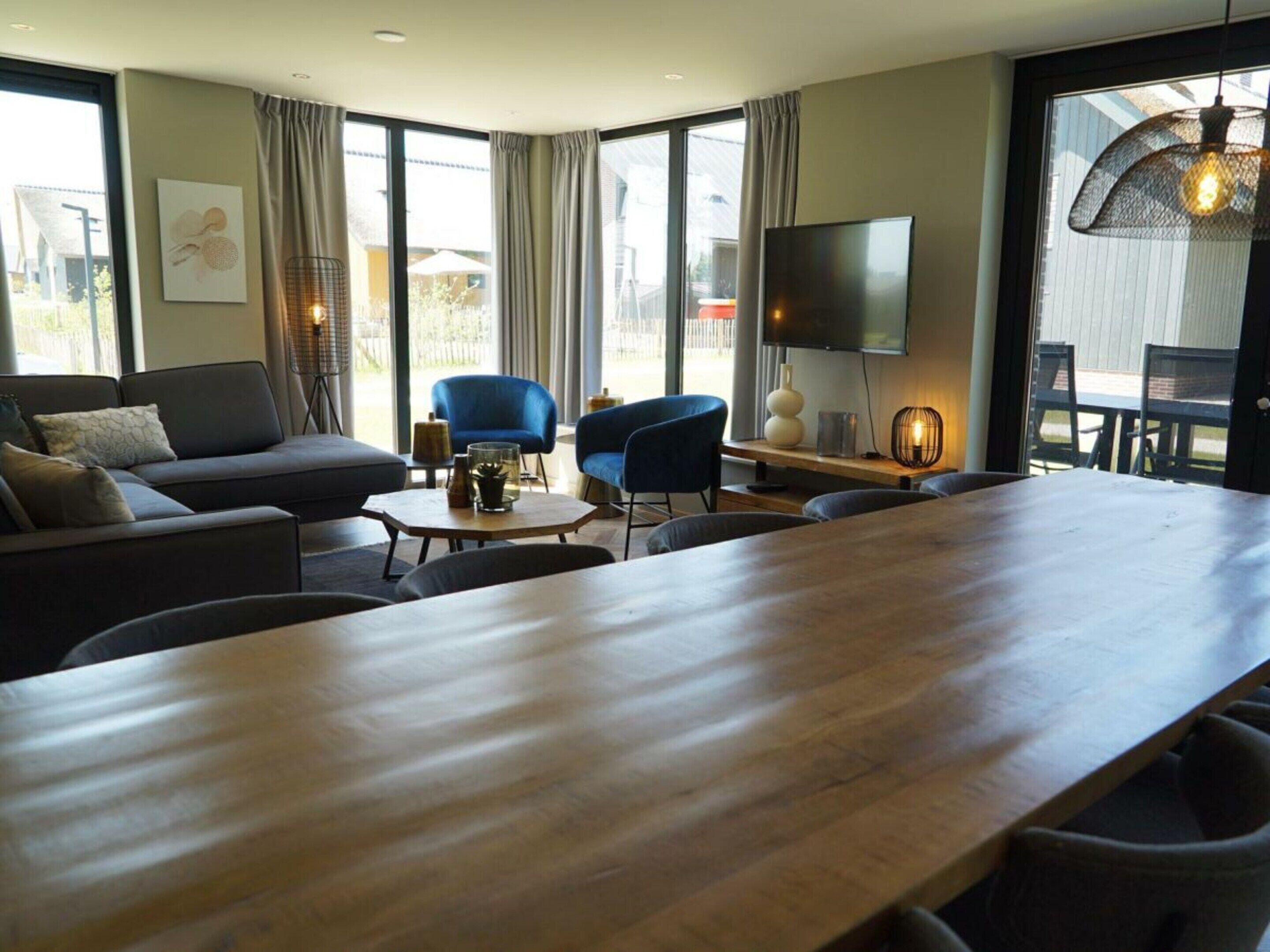 Un salon moderne avec une longue table en bois, un canapé, deux fauteuils bleus, un téléviseur et de grandes fenêtres avec des rideaux.