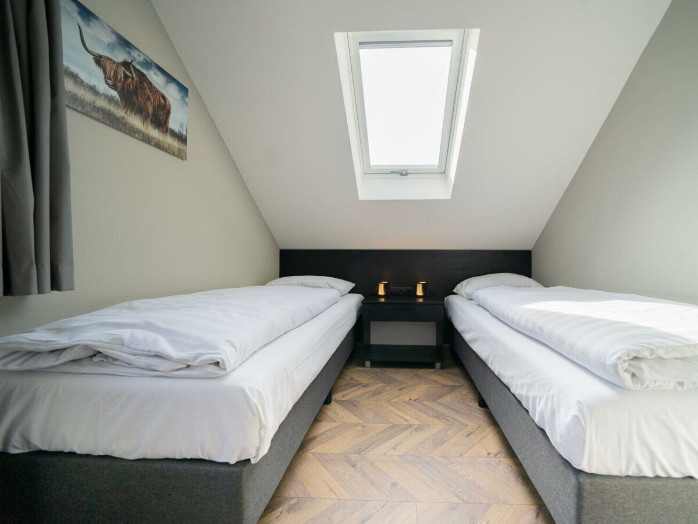 A small, modern bedroom with two single beds, a skylight, a herringbone floor, and a painting on the wall.