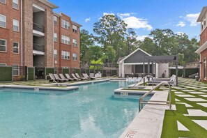 A swimming pool with lounge chairs, a covered seating area, and a brick building in the background. - NEW Mary Harmon Landing 1st Floor w/ Gym & Pool (Tuscaloosa)