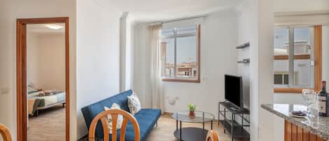 A modern living room with a blue sofa, a dining table with fruit, a TV, and a kitchen area with a wine bottle.