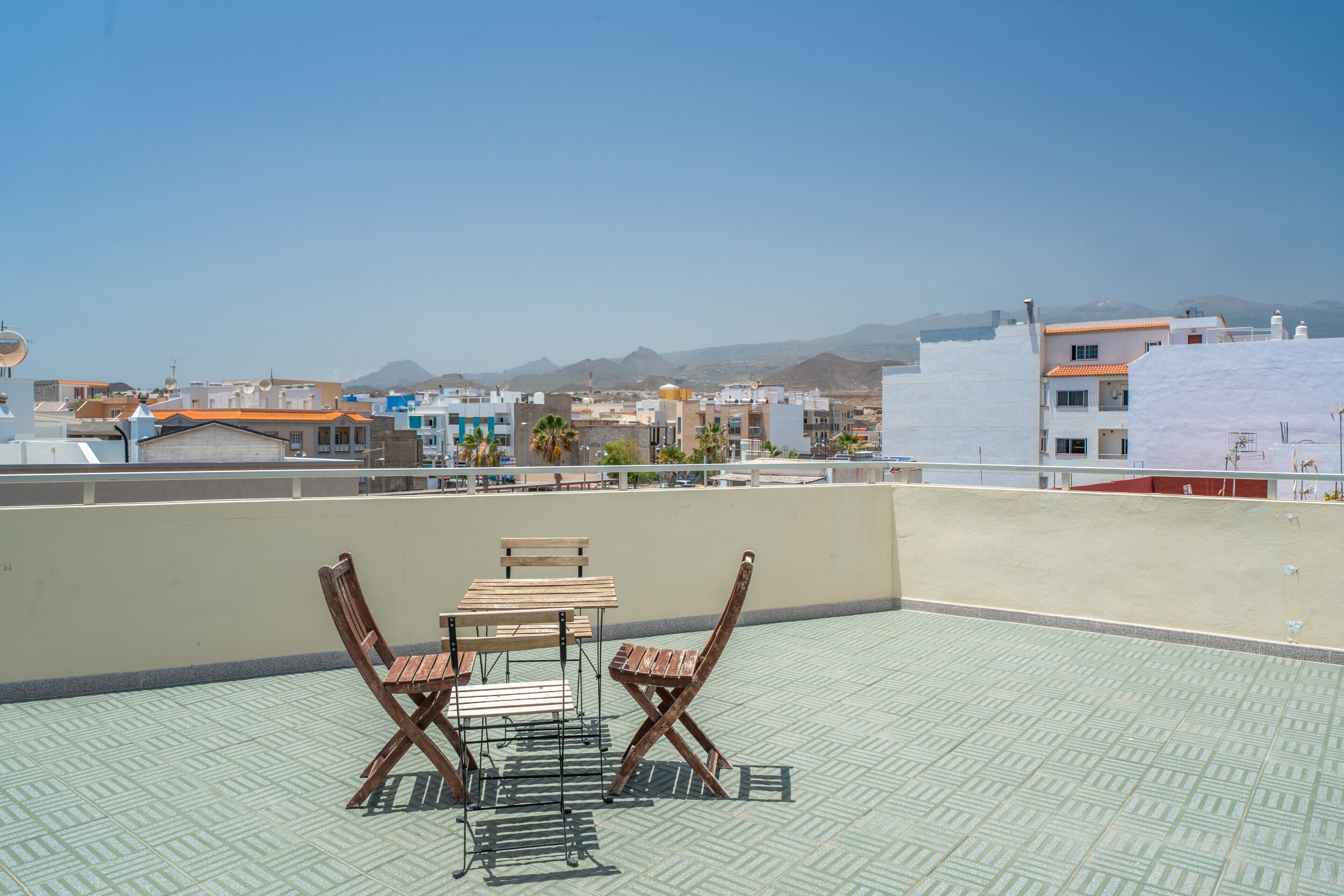 Una terraza en la azotea con una mesa y sillas, suelo de baldosas y vistas a edificios y montañas a lo lejos.