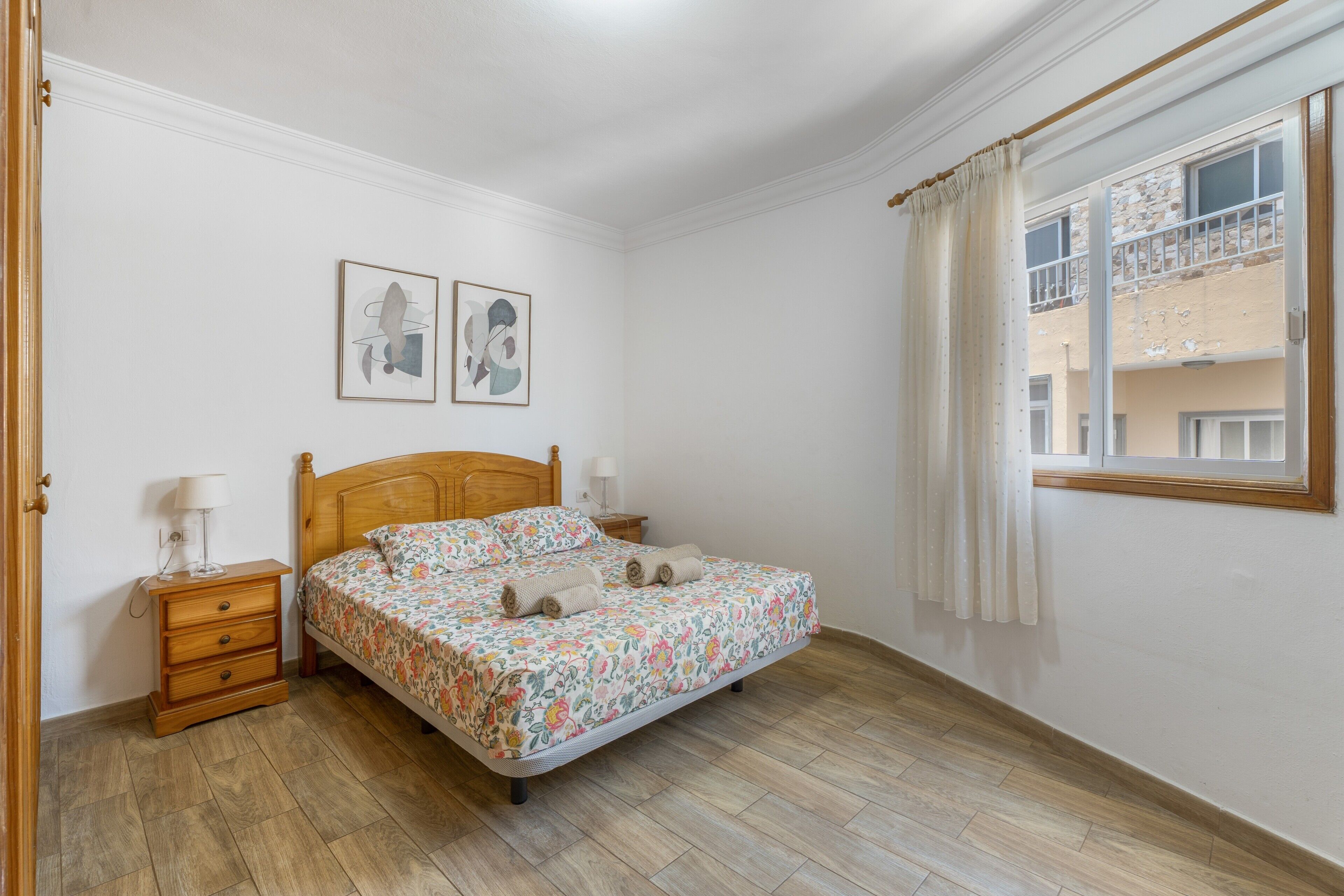 Une chambre à coucher avec un lit en bois, une table de chevet, une fenêtre avec des rideaux et deux tableaux accrochés au mur.