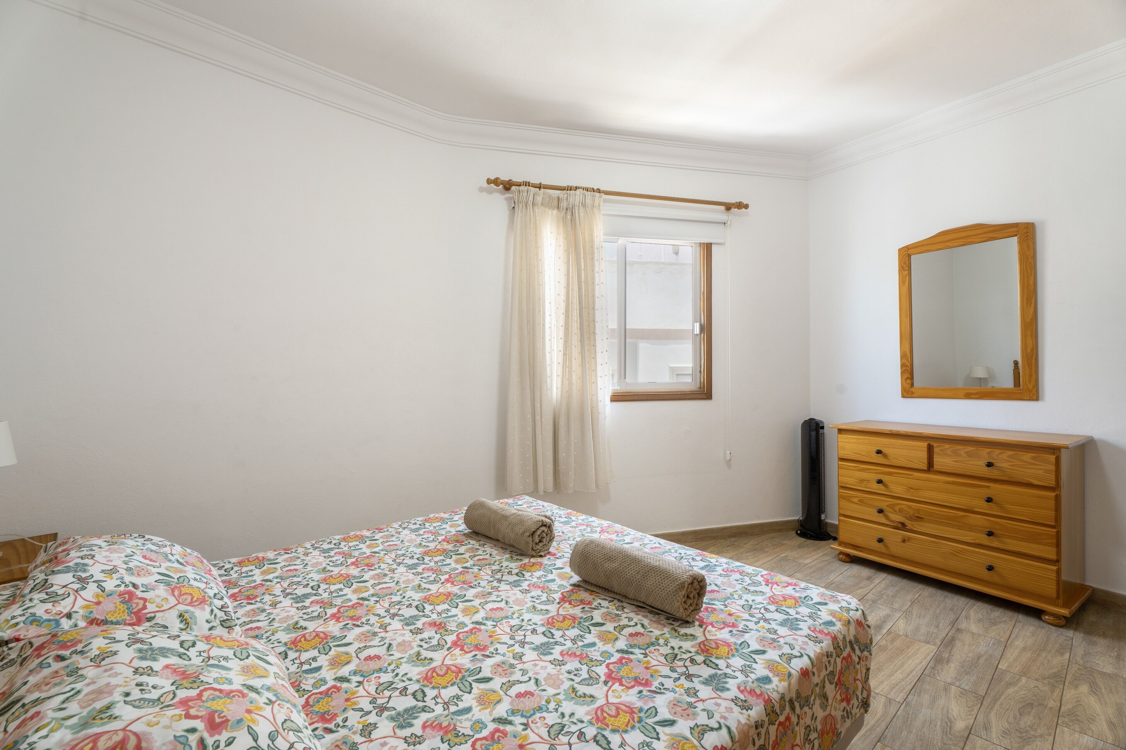 Een slaapkamer met een bed, een dressoir, een spiegel en een raam met gordijnen.