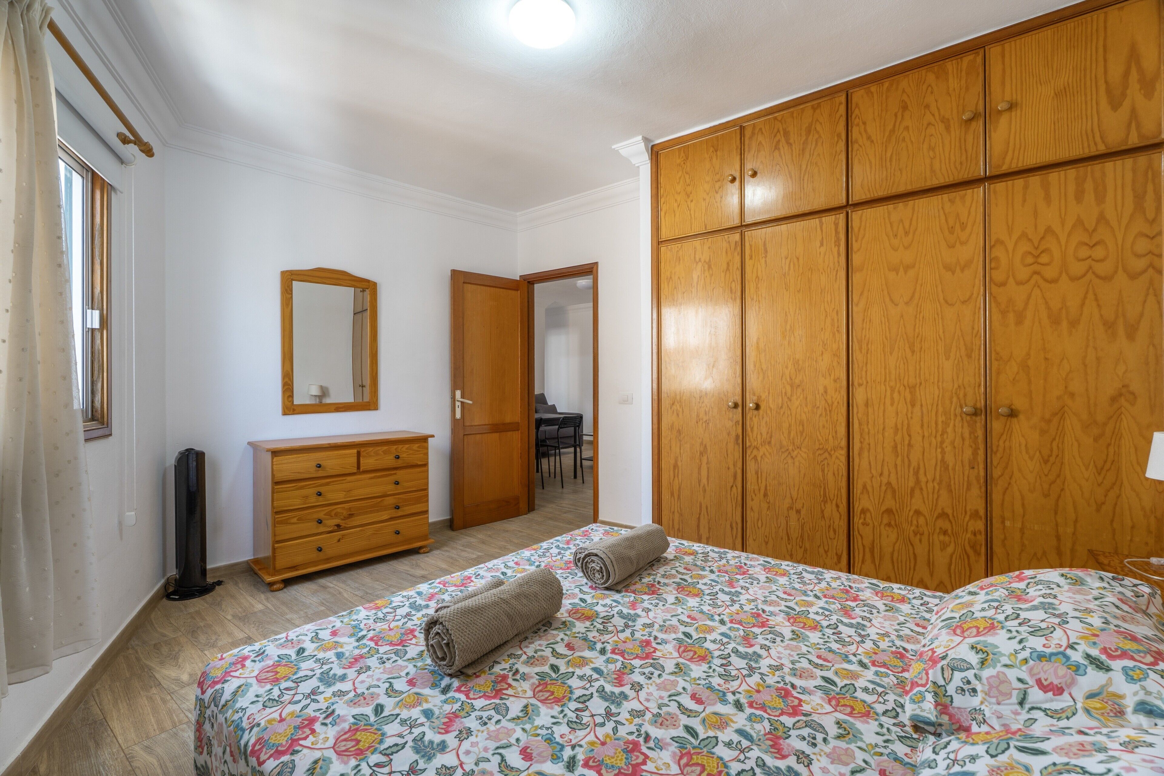A bedroom with a bed, a wooden wardrobe, a dresser, a mirror, and a door leading to another room.