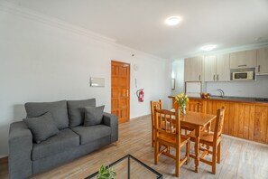 Ein Wohnzimmer mit grauem Sofa, einem Holzespeisetisch und Stühlen. Im Hintergrund ist eine Küche mit Holzschränken und einer Mikrowelle zu sehen.