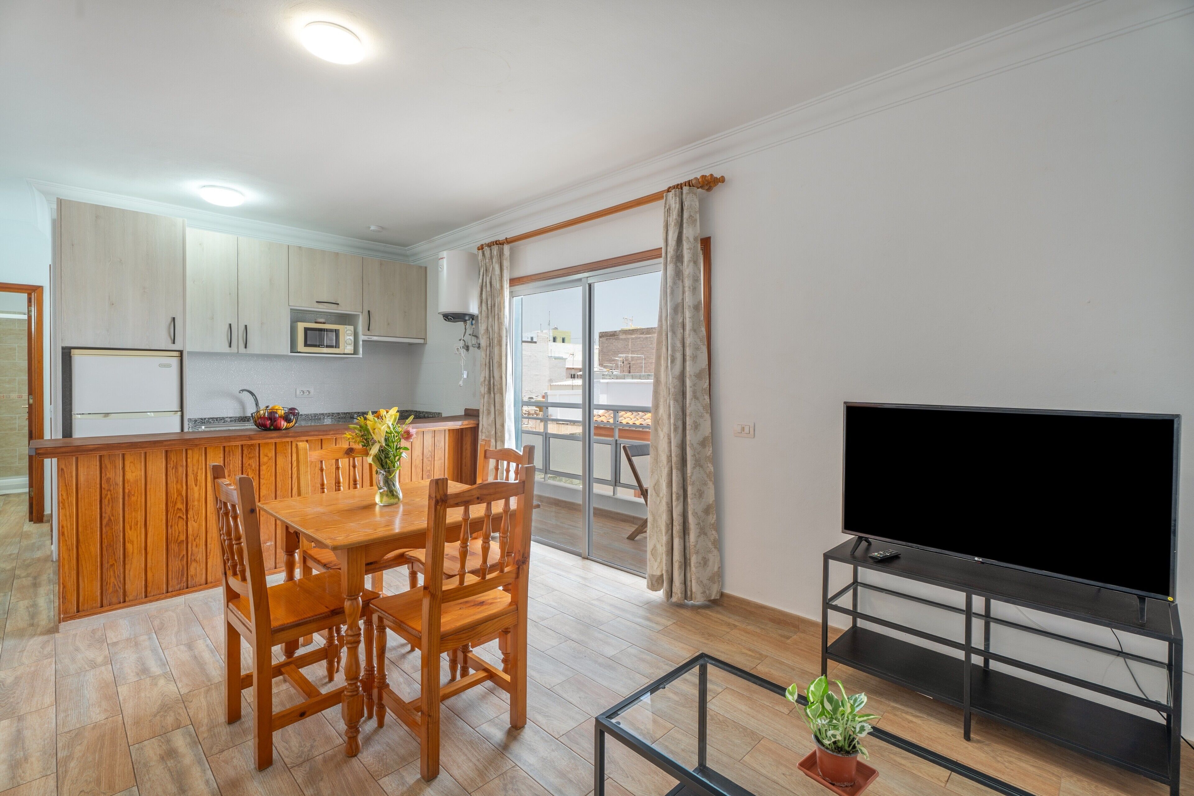 Una cocina moderna con armarios de madera, una mesa de comedor con sillas, un microondas empotrado y un televisor sobre un mueble.