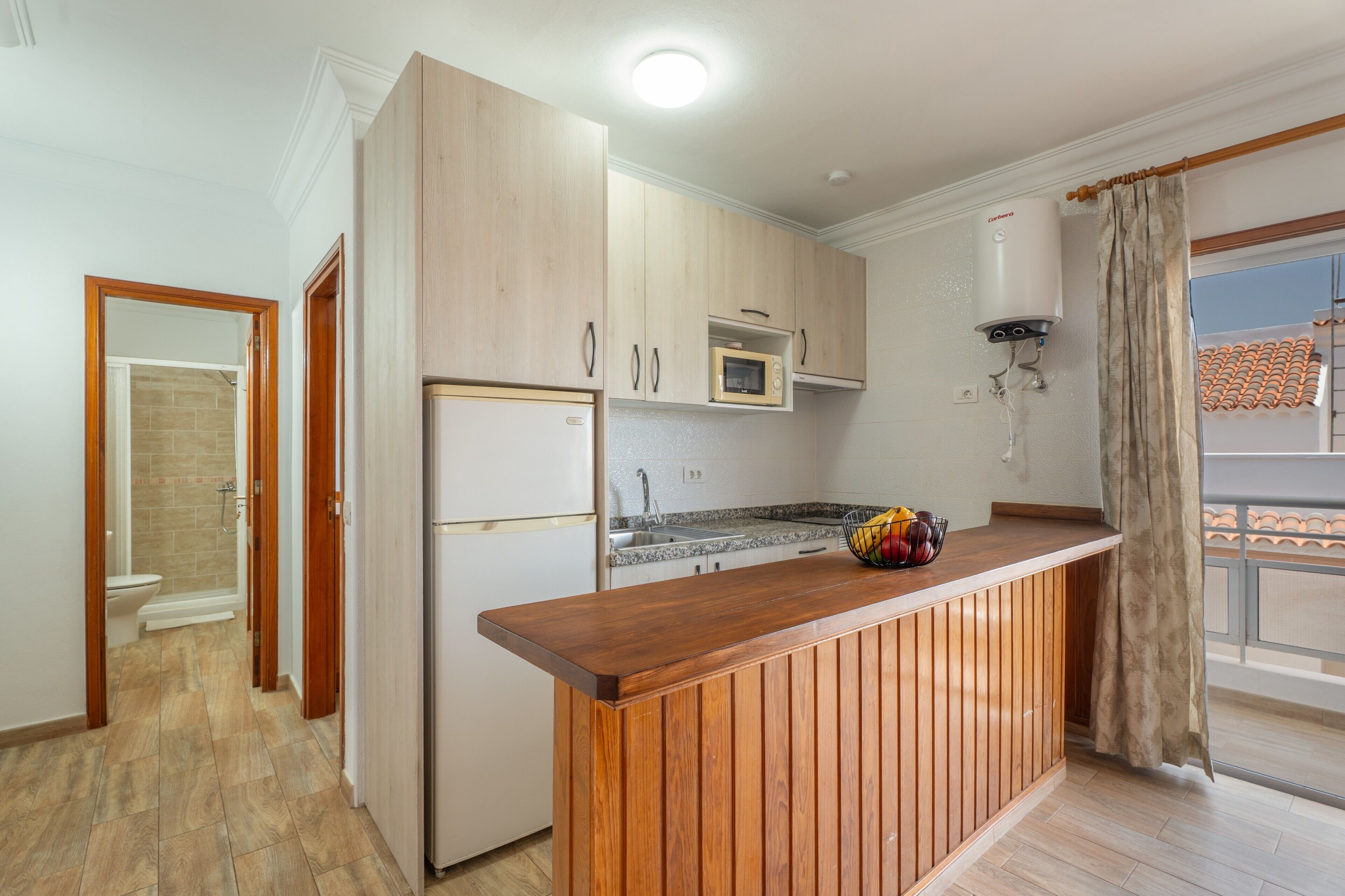Een compacte keuken met houten kastjes, een inbouwmagnetron en een klein eiland met een schaal fruit.