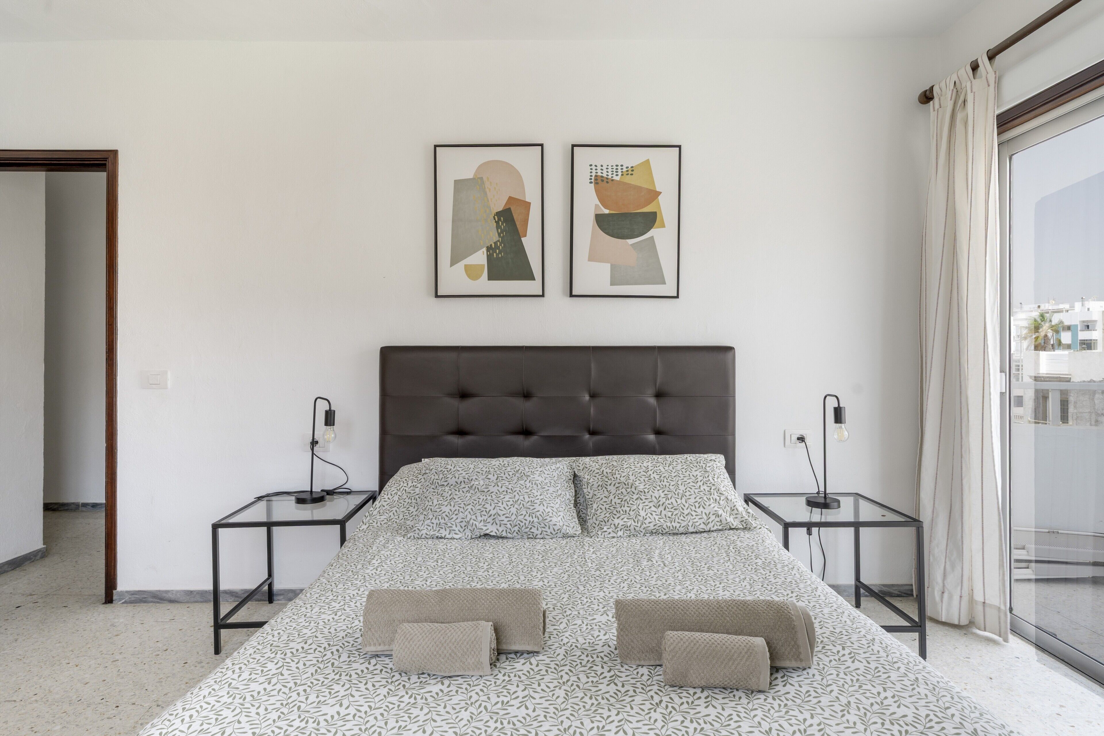 Un dormitorio moderno con un cabecero capitonado, una cama grande con ropa de cama estampada, dos mesitas de noche con lámparas y una obra de arte abstracta en la pared.