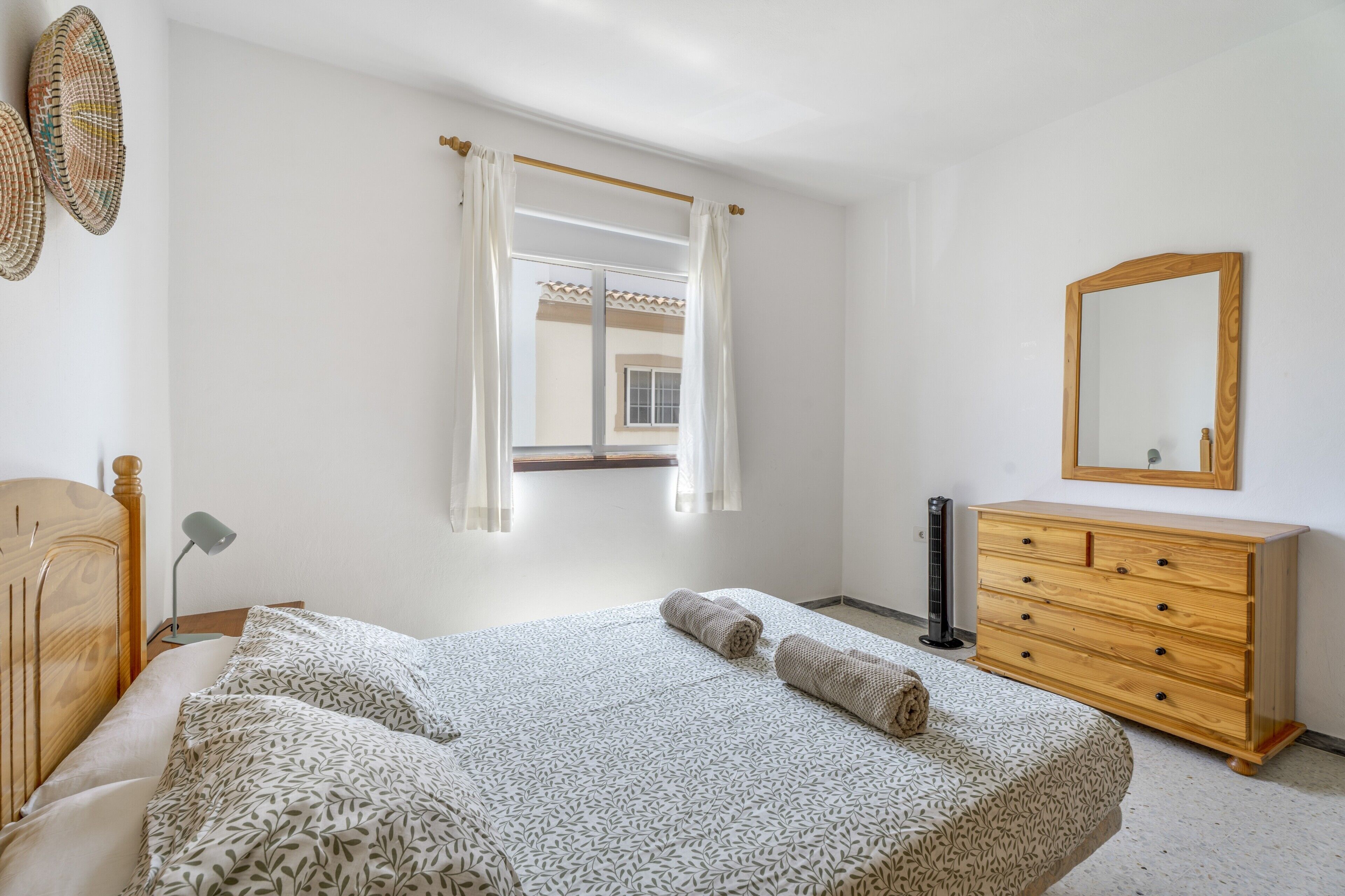 Een slaapkamer met een bed, een houten dressoir, een spiegel en een raam met gordijnen.