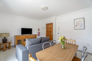 Ein modernes Wohnzimmer mit Flachbildfernseher, grauem Sofa, Esstisch aus Holz und einer Blumenvase.