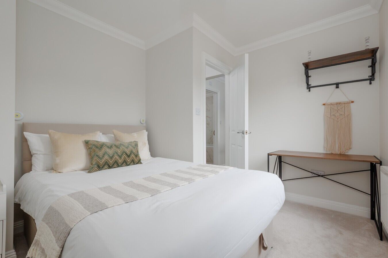 A bedroom with a bed, a bedside table, a wall-mounted shelf, and a door leading to another room.