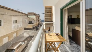 Un balcón con una mesa, una botella de vino y vasos, con vista a una calle con coches aparcados y edificios.