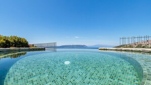 Ein klarer, blauer Infinity-Pool mit gefliestem Beckenboden, umgeben von einer niedrigen Mauer und einem Metallzaun, mit Blick auf eine entfernte Küstenlinie.