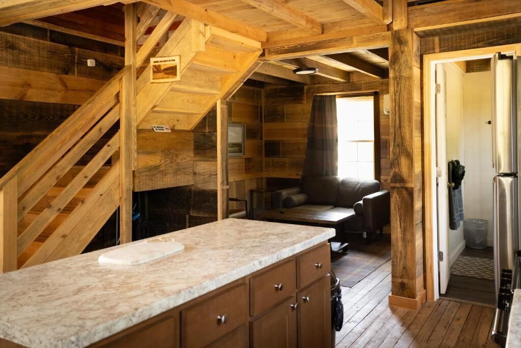 A rustic cabin interior with a kitchen area, wooden walls, a staircase, and a cozy seating area.