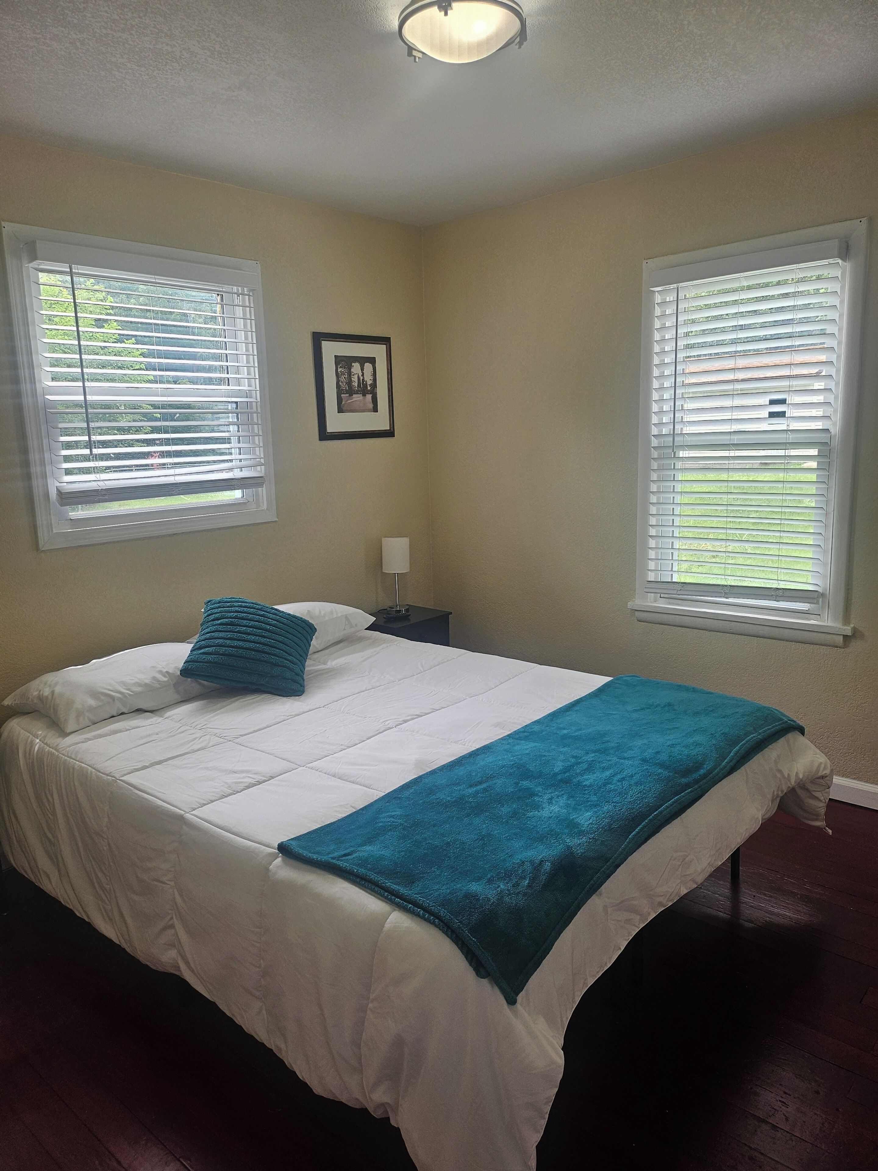 A bedroom with a bed, two windows with blinds, a nightstand, a lamp, and a framed picture on the wall.