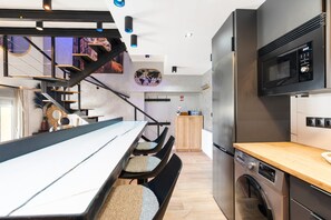 Een moderne keuken met een magnetron, oven en wasmachine. Er is een eethoek met een lange tafel en stoelen. Een trap leidt naar een verdieping.