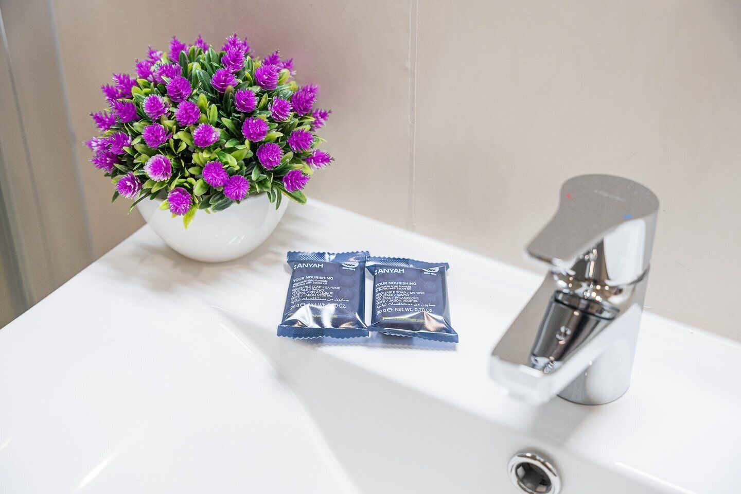 A bathroom sink with a white basin, a potted plant with purple flowers, and two packets of soap.