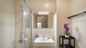 Un baño con cabina de ducha de vidrio, un lavamanos blanco, un espejo sobre el lavamanos y una repisa con una orquídea en maceta.