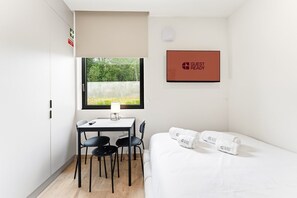 Ein Zimmer mit einem Bett, einem Schreibtisch mit zwei Stühlen, einem Fenster mit Blick ins Grüne und einem gerahmten Schild an der Wand.