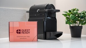 Una cafetera negra, una caja rosa con la inscripción 'GUEST READY' y una planta en maceta sobre una superficie blanca.