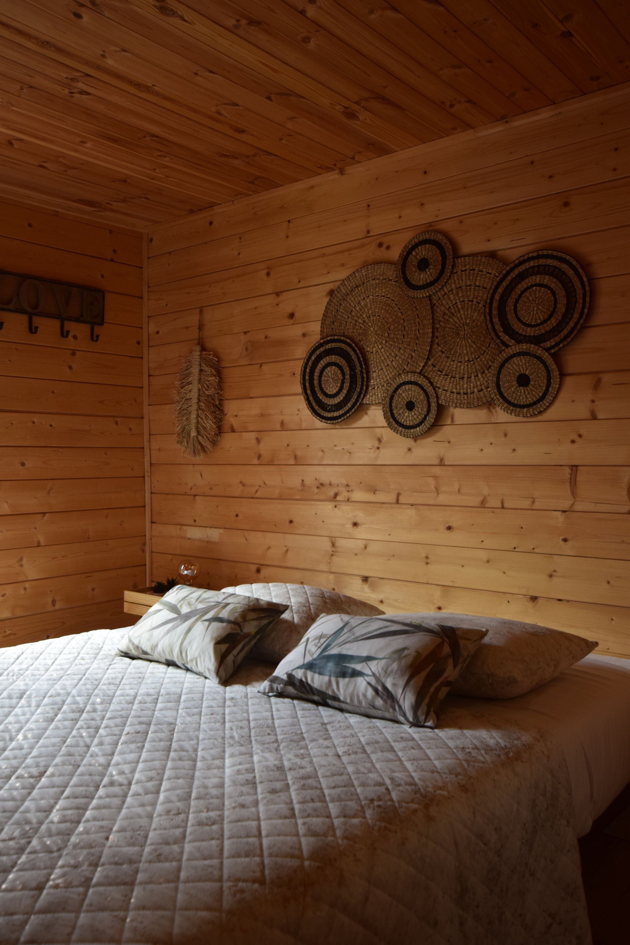 A wooden cabin interior with a bed, a feather wall hanging, and woven wall decorations.