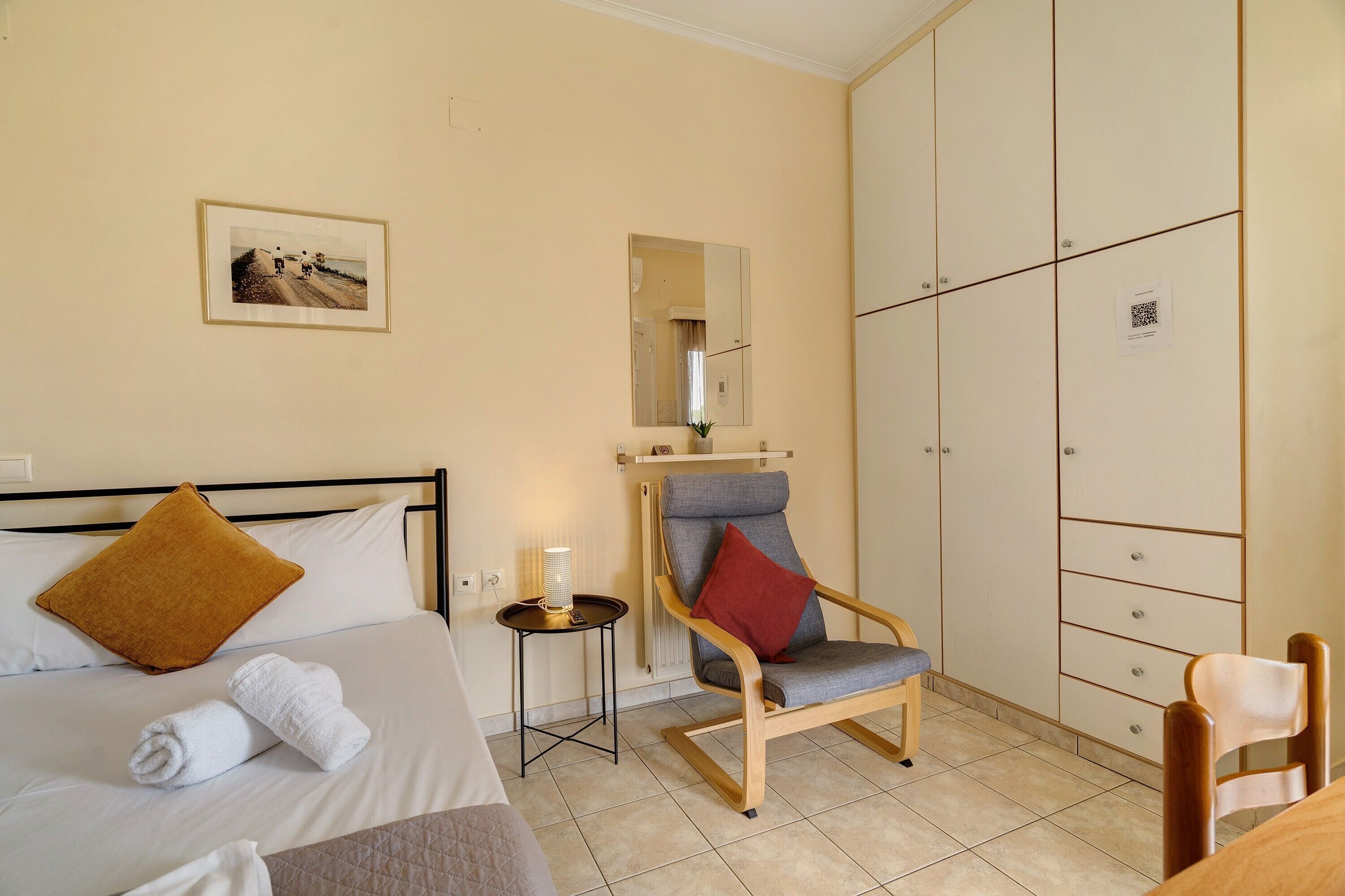 A hotel room with a bed, a chair, a small table with a lamp, a wardrobe, and a painting on the wall.